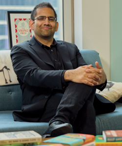 Ibrahim Ahmad, a man of Middle Eastern descent with tan skin, glasses, and short, black hair sits with his hands folded and legs crossed in front of a table of books.