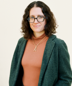 Janelle Bassett, a white woman with glasses and brown, wavy hair, stands with her hands in her pockets and wears a burnt orange turtleneck with an emerald green blazer.